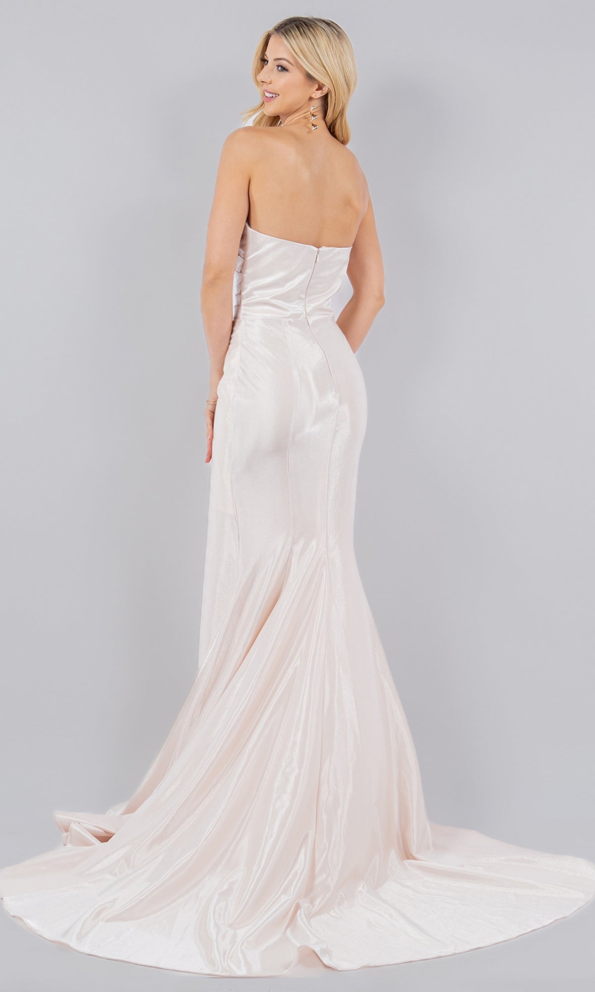 A blonde woman poses with her back to the camera in the Cinderella Couture 8083J, a strapless, floor-length light pink satin mermaid prom dress with a slight train, set against a gray background.